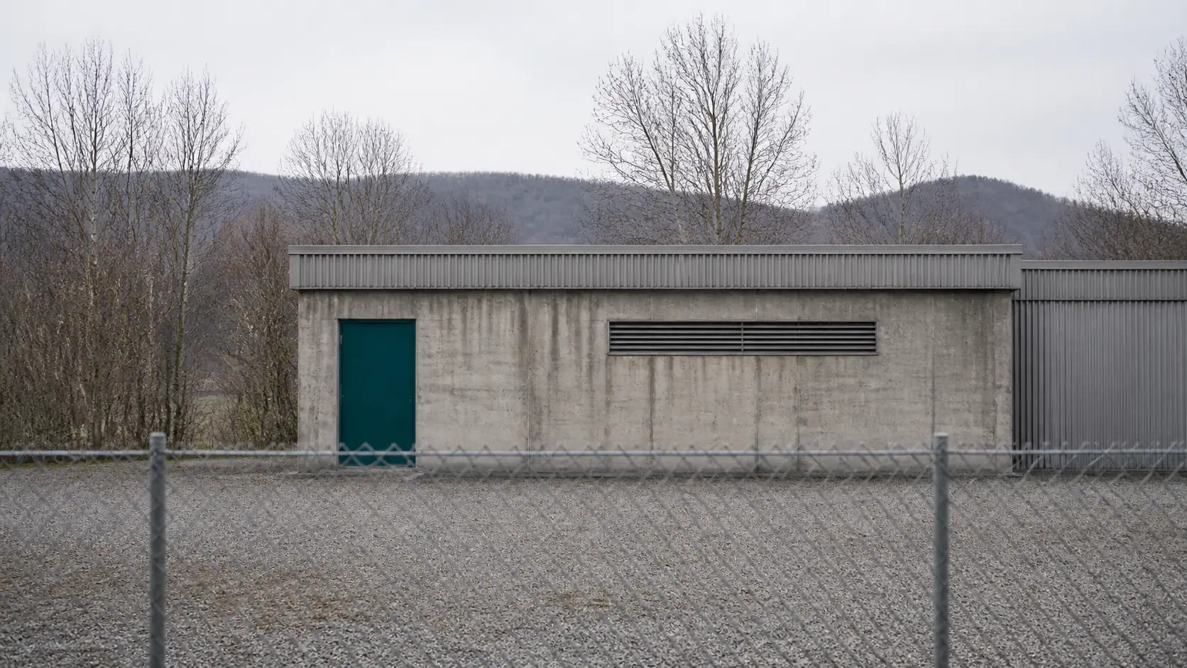 Ein niedriges fensterloses Zweckgebäude aus Beton hinter einem Maschendrahtzaun, dahinter bewaldete Hügel